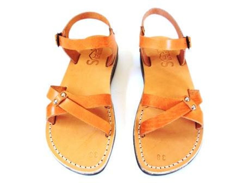 May include: A pair of tan leather sandals with a cross-strap design and ankle straps. The sandals have a stitched sole and a buckle closure. The sandals are size 38. The sandals are displayed on a white background.