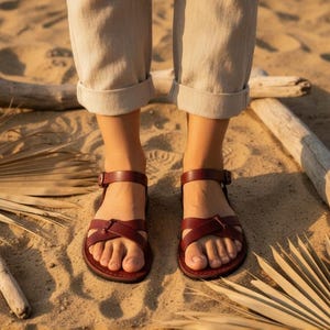 Dark brown minimalist leather sandals on a sandy beach. Rustic flat summer shoes paired with linen trousers for a natural organic look.