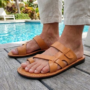 Puede incluir: Sandalias de cuero color canela con suela negra. Las sandalias tienen una presilla para el dedo gordo y múltiples correas en la parte superior del pie. Llevadas en una terraza de madera cerca de una piscina.