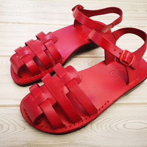 Pode incluir: Um par de sand&aacute;lias de couro vermelho com um design de tira tecida. As sand&aacute;lias t&ecirc;m um fecho de fivela e uma sola plana.