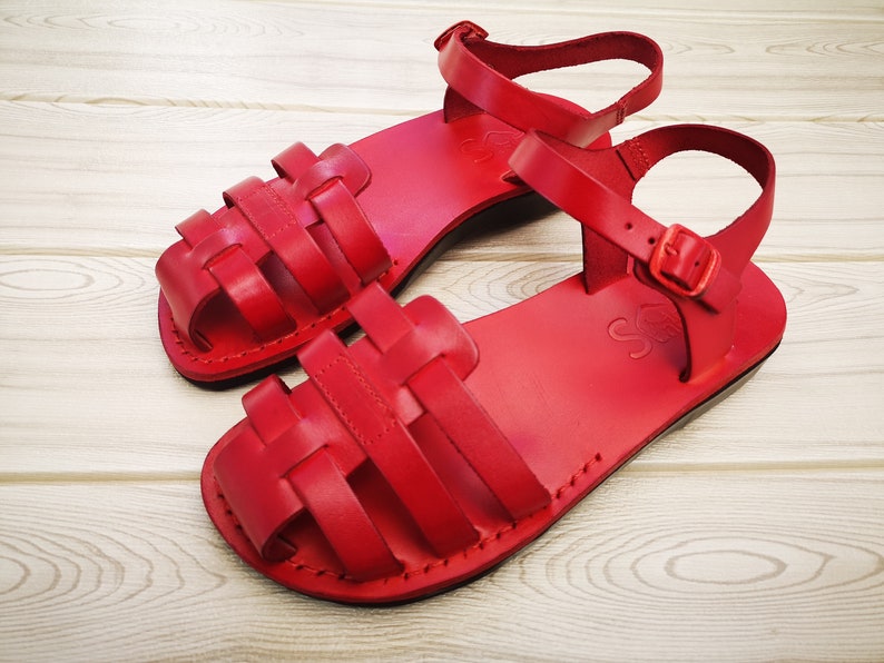 Pode incluir: Um par de sand&aacute;lias de couro vermelho com um design de tira tecida. As sand&aacute;lias t&ecirc;m um fecho de fivela e uma sola plana.