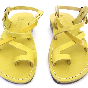 K&ouml;nnte beinhalten: Ein Paar gelbe Ledersandalen mit gekreuzten Riemen und einer Zehenschlaufe. Die Sandalen haben einen Schnallenverschluss am Kn&ouml;chelriemen.