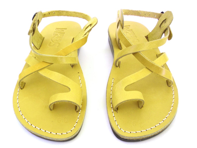 K&ouml;nnte beinhalten: Ein Paar gelbe Ledersandalen mit gekreuzten Riemen und einer Zehenschlaufe. Die Sandalen haben einen Schnallenverschluss am Kn&ouml;chelriemen.