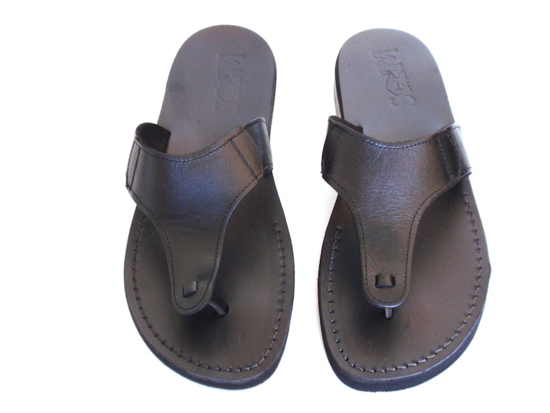 Peut inclure: Une paire de sandales en cuir noir &agrave; lani&egrave;res avec un design simple. Les sandales ont un bord cousu et un petit d&eacute;tail carr&eacute; sur la lani&egrave;re.
