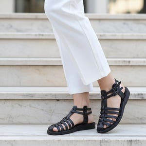 Elegant black leather gladiator sandals walking down marble stairs. Classic women's handmade summer shoes offering chic comfort for formal or casual wear.
