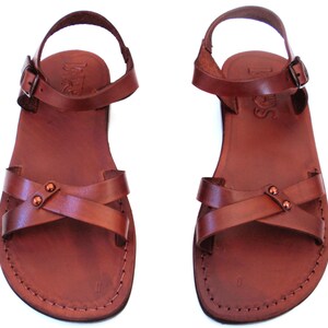 May include: A pair of brown leather sandals with a cross-strap design and adjustable ankle straps. The sandals have a stitched sole and a buckle closure. The leather appears to be a rich, reddish-brown color.