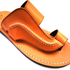 May include: A pair of tan leather sandals with white stitching. The sandals have a simple design with a single strap across the top of the foot and a toe loop.