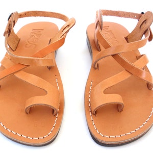 K&ouml;nnte beinhalten: Tannbraune Ledersandalen mit gekreuzten Riemen und Zehenschlaufe. Die Sandalen haben einen Schnallenverschluss am Kn&ouml;chel und der Markenname "INUOVO" ist auf den Riemen gepr&auml;gt.