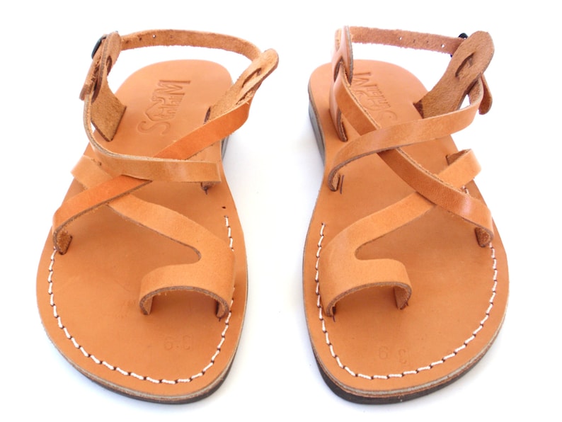 K&ouml;nnte beinhalten: Tannbraune Ledersandalen mit gekreuzten Riemen und Zehenschlaufe. Die Sandalen haben einen Schnallenverschluss am Kn&ouml;chel und der Markenname "INUOVO" ist auf den Riemen gepr&auml;gt.