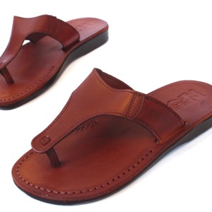 Peut inclure: Une paire de sandales en cuir marron avec une simple lani&egrave;re et un bord cousu. Les sandales ont une semelle plate et confortable.