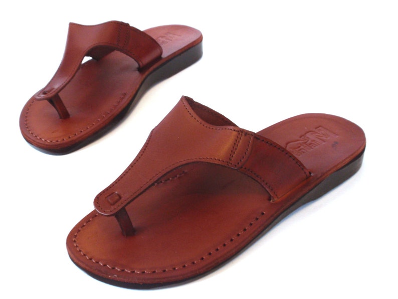 Peut inclure: Une paire de sandales en cuir marron avec une simple lani&egrave;re et un bord cousu. Les sandales ont une semelle plate et confortable.