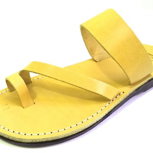 May include: A pair of yellow leather sandals with a simple design. The sandals have a single strap that goes around the ankle and a toe loop. The sole is made of leather and has a stitched edge.