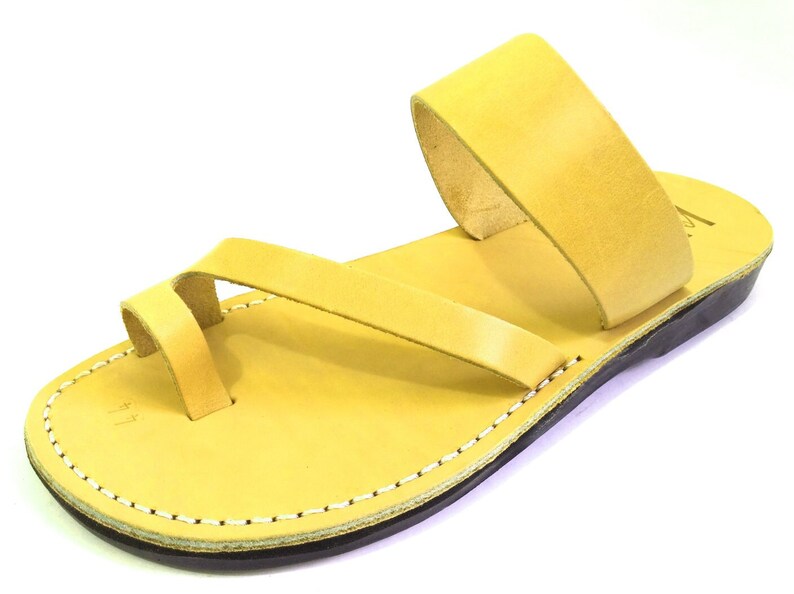 May include: A pair of yellow leather sandals with a simple design. The sandals have a single strap that goes around the ankle and a toe loop. The sole is made of leather and has a stitched edge.
