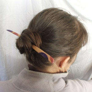 May include: A wooden hair stick with purple, red and white painted circles. The stick is being used to hold a woman's hair in a bun.