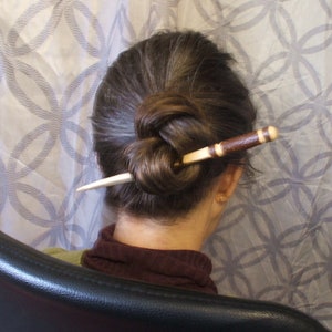 May include: A brown and white wooden hair stick with a knot at the end. The stick is being used to hold a woman's brown hair in a bun.