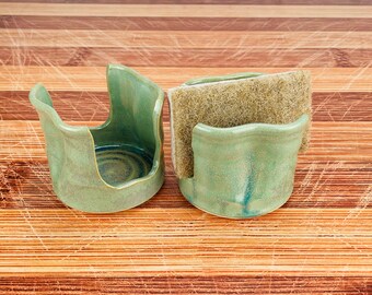 May include: Two handmade ceramic sponge holders in a seafoam green glaze. One holder is empty, the other holds a tan scrub sponge. The holders have a curved design and sit on a wooden surface.