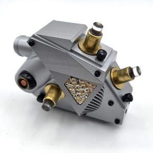 May include: A silver-toned mechanical device with a geometric, angular body. It features two brass-colored cylindrical protrusions topped with clear glass, a triangular inset filled with small metallic circles, and several black screws.