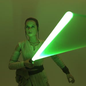 Puede incluir: Figura de Rey de Star Wars empuñando un sable de luz verde. El sable de luz emite un brillo verde brillante, contrastando con el atuendo beige de la figura y el fondo tenue. La figura está en pose de batalla.