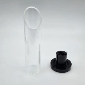 Op de afbeelding: Een heldere, cilindrische glazen buis met een schuine bovenkant, naast een zwarte, cirkelvormige basis met een centrale paal. De basis heeft een sleuf. De objecten staan op een witte achtergrond.