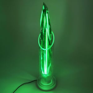 Op de afbeelding: Een groene neonlichtsculptuur in een heldere glazen cilinder. De sculptuur heeft een puntige top en een cirkelvormig neonaccent. De basis is wit en een zwart snoer loopt van de onderkant. Het algehele effect is een gloeiend, modern kunstwerk.