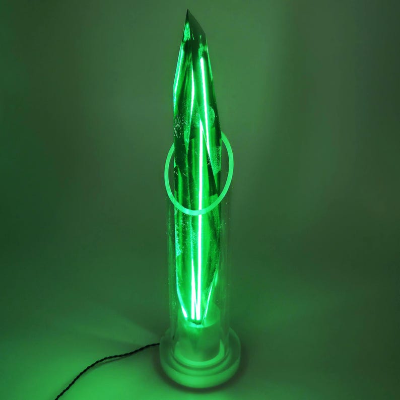 Op de afbeelding: Een groene neonlichtsculptuur in een heldere glazen cilinder. De sculptuur heeft een puntige top en een cirkelvormig neonaccent. De basis is wit en een zwart snoer loopt van de onderkant. Het algehele effect is een gloeiend, modern kunstwerk.
