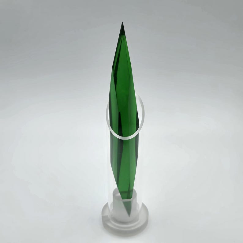Op de afbeelding: Een hoge, groene, geometrische sculptuur met een puntige top, tentoongesteld in een heldere, cilindrische houder. De sculptuur heeft een reeks donkergroene en zwarte verticale lijnen. Een heldere, cirkelvormige ring omringt de sculptuur.