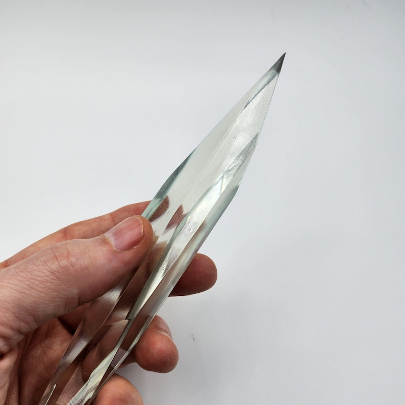K&ouml;nnte beinhalten: Eine klare, facettierte Kristallspitze mit einer scharfen Spitze. Der Kristall wird in einer Hand gehalten, wodurch seine L&auml;nge und Form hervorgehoben werden.