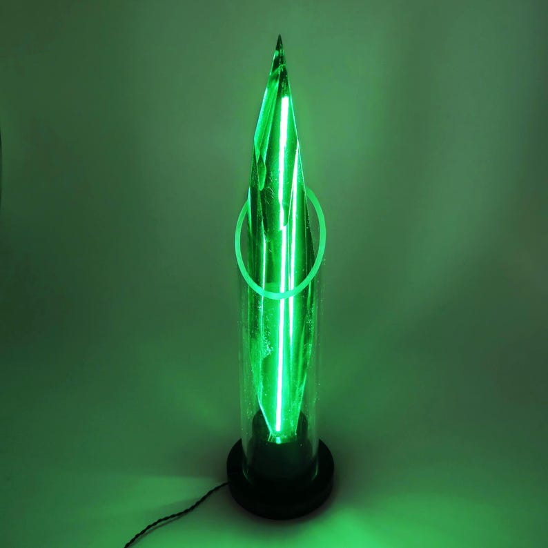 Op de afbeelding: Een groene, verlichte sculptuur die lijkt op een kristal, ingesloten in een heldere cilindrische buis. De sculptuur heeft een puntige top en wordt verlicht met heldergroene neonlichten. De basis is zwart en er loopt een netsnoer uit.