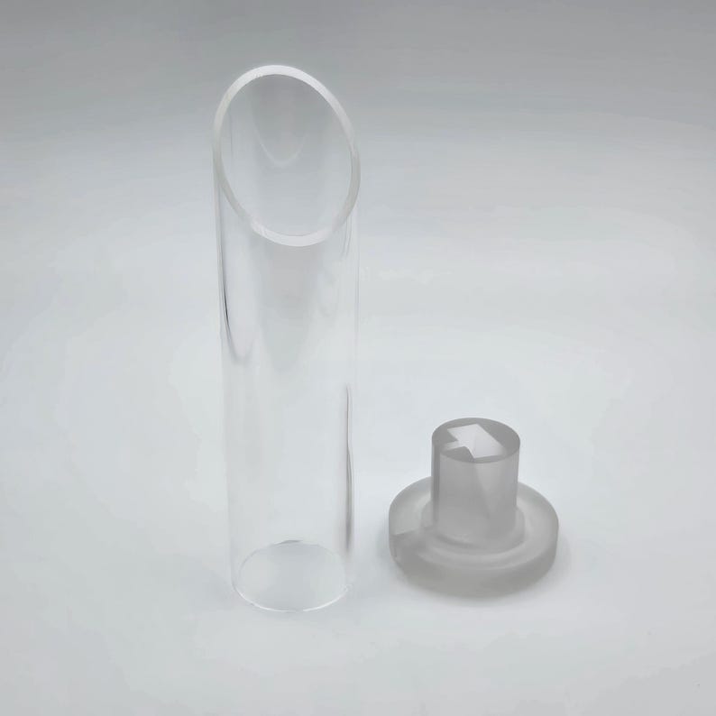 Op de afbeelding: Een heldere, cilindrische glazen buis met een schuine bovenkant, naast een matglazen stopper. De stopper heeft een cirkelvormige basis en een cilindrische bovenkant met een vierkant ontwerp. De objecten staan op een witte achtergrond.