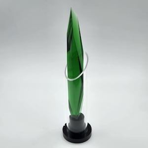 Op de afbeelding: Een hoge, groene glazen sculptuur met een puntige top, omhuld door een heldere cilinder. Een zilveren ring omcirkelt de sculptuur. De basis is zwart. De sculptuur heeft een modern, geometrisch ontwerp.