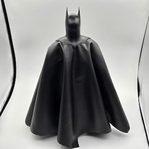 Puede incluir: Una figura negra de Batman, con capucha y capa. La figura está de pie, con la capa extendida detrás. La figura está hecha de un material liso y mate.