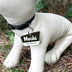 May include: A white plush dog toy with a black collar and a brown tag that says "Mochi" and a phone number. The toy is sitting on a bed of pine needles and grass.