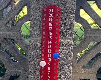May include: A red wooden bean bag game scoreboard with white and blue scoring pegs. The scoreboard is labeled "THE SARLOS" and has numbers 0-21. The game is set up on a wooden platform with blue and white star-patterned bean bags.