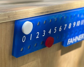 May include: A blue plastic scoreboard with white numbers 0-10 and a red and white counter. The scoreboard is attached to a wooden surface.