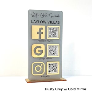 May include: A dusty grey sign with a gold mirror finish. The sign has the text "Let's Get Social" and "Laylow Villas" in black lettering. There are three social media icons with QR codes: Facebook, Google, and Instagram.