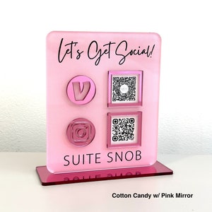 May include: A pink acrylic social media display stand with the text "Let's Get Social!" and the brand name "SUITE SNOB". The stand features icons for social media platforms and QR codes. The base is a darker pink mirror.