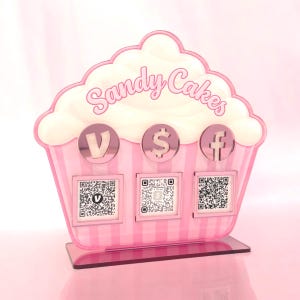 May include: A cupcake-shaped acrylic sign in pink and white with the text Sandy Cakes. It features three circular icons for Venmo, Cash App, and Facebook, each positioned above a corresponding square QR code on a flat rectangular base.