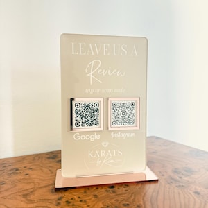 May include: A rectangular acrylic sign with a rose gold base featuring the text LEAVE US A Review tap or scan code. It includes two square QR codes with rose gold frames labeled Google and Instagram, and the logo KARATS by Kim at the bottom.