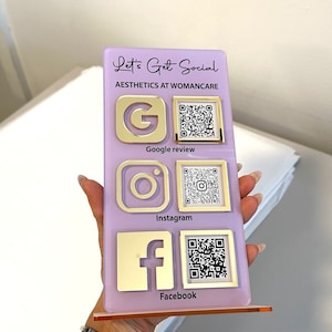 Op de afbeelding: Een paars bord met de tekst "Let's Get Social" en "Aesthetics at Womancare" in gouden letters. Het bord heeft drie sociale media-iconen: Google, Instagram en Facebook, elk met een bijbehorende QR-code.
