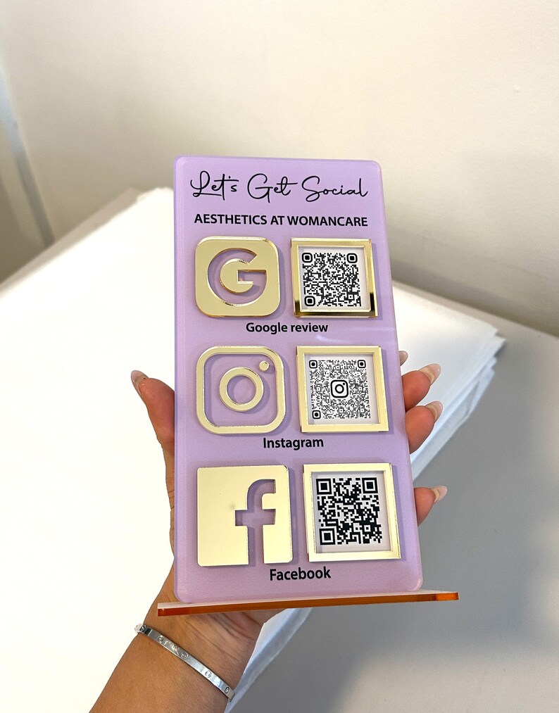 Op de afbeelding: Een paars bord met de tekst "Let's Get Social" en "Aesthetics at Womancare" in gouden letters. Het bord heeft drie sociale media-iconen: Google, Instagram en Facebook, elk met een bijbehorende QR-code.