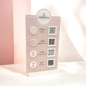 May include: A light pink acrylic sign with a white base featuring the Simply Baked Goods by Debrise logo, four white social media and payment icons, corresponding text labels, and four square QR codes for Instagram, website, CashApp, and Zelle.