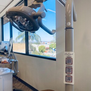 May include: A white dental light fixture with a gray arm attached to a white pole. The pole has a white sticker with the text "Let's Connect" and QR codes for Google, Instagram, and a menu.