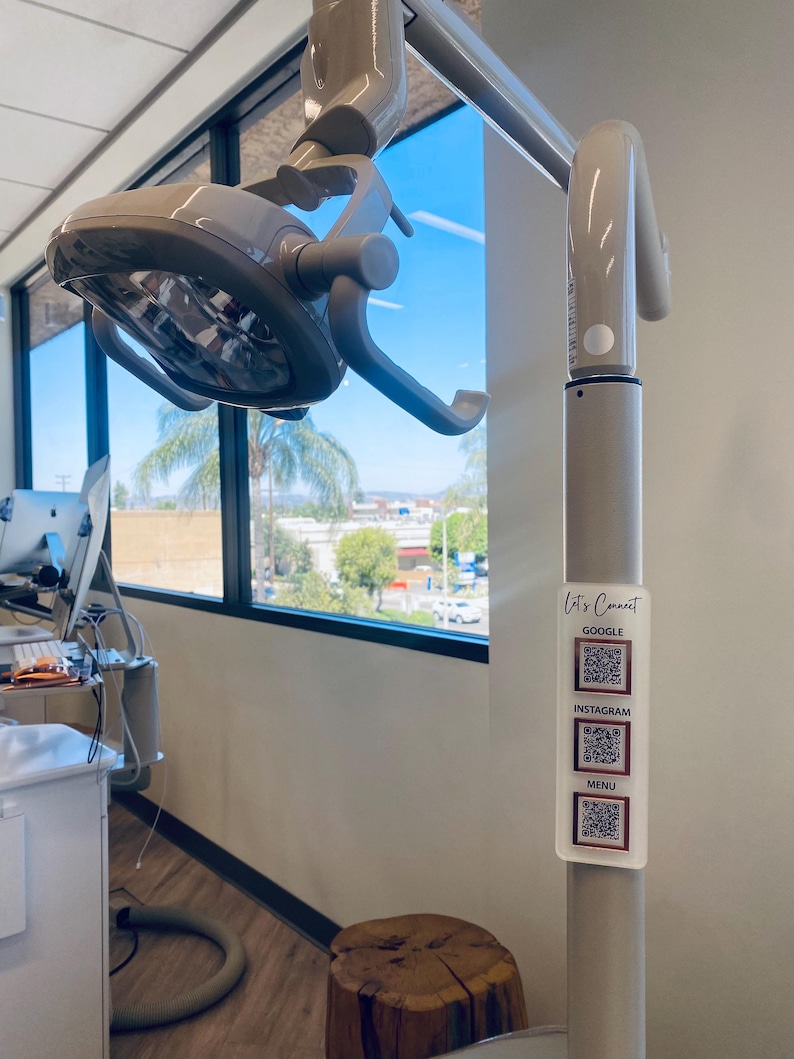 May include: A white dental light fixture with a gray arm attached to a white pole. The pole has a white sticker with the text "Let's Connect" and QR codes for Google, Instagram, and a menu.