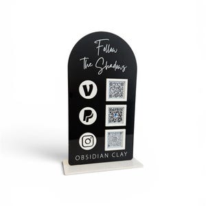 May include: A black acrylic display with a curved top and a white base. The display features the text "Follow The Shadows" in cursive, along with social media icons and QR codes. The text "OBSIDIAN CLAY" is at the bottom.
