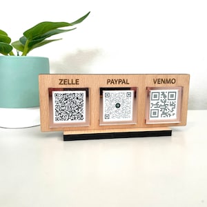 May include: A wooden stand with three separate sections, each with a different payment method: Zelle, PayPal, and Venmo. Each section has a QR code printed on a white background.