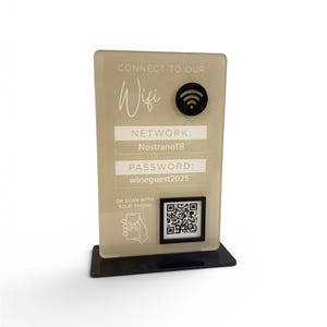 May include: A rectangular, beige-colored WiFi sign with black text and a black base. The sign displays the text "CONNECT TO OUR Wifi", network name "NostranoTR", and password "wineguest2025". A QR code is also present.