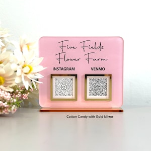 May include: Pink acrylic sign with the text "Five Fields Flower Farm" and two QR codes labeled "Instagram" and "Venmo". The sign is on a wooden stand and has a gold mirror finish. The sign is in front of a bouquet of white flowers.