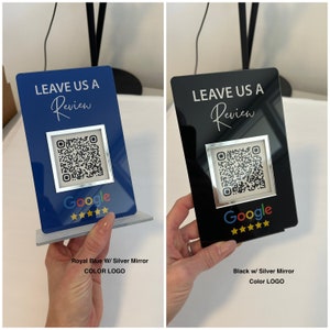 K&ouml;nnte beinhalten: K&ouml;nigsblaue und schwarze Bewertungsschilder mit silbernen Spiegelakzenten. Jedes Schild zeigt einen QR-Code, das Google-Logo und den Text "LEAVE US A Review". Das k&ouml;nigsblaue Schild hat einen transparenten Sockel, w&auml;hrend das schwarze Schild von einer Hand gehalten wird.