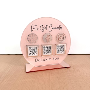 May include: A pink acrylic stand with the text "Let's Get Connected" and the logos for Facebook, Instagram, and a globe. There are three QR codes below the logos and the text "DeLuxie Spa" at the bottom of the stand.