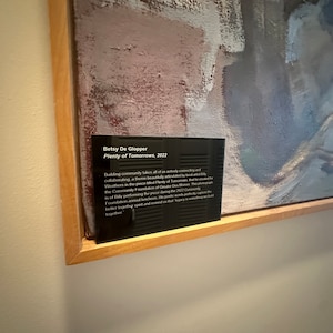 May include: A black plaque with white text on a wooden frame. The text reads "Betsy De Glopper, Plenty of Tomorrows, 2022". The plaque is mounted on a wall next to an abstract painting with shades of gray, pink, and white.